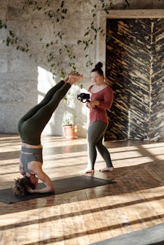 Woman practicing headstand yoga pose indoors, with another woman photographing her.