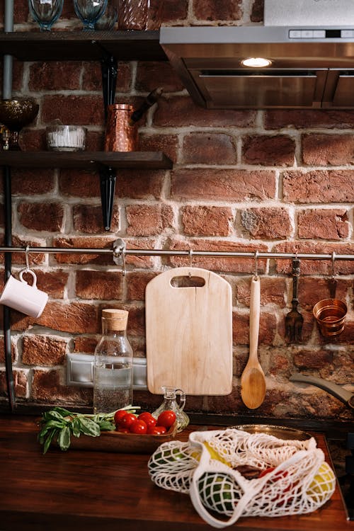 Rustic kitchen corner with a cutting board, fresh vegetables, and cooking utensils against a brick wall.
