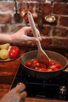 A person cooking cherry tomatoes with a wooden spoon on a stovetop in a cozy kitchen setting.