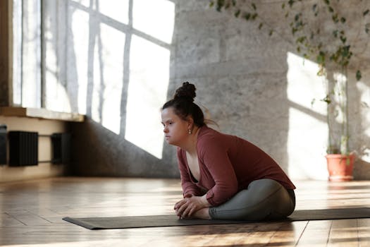 A woman with Down syndrome sits on a yoga mat indoors in a relaxed yoga pose.