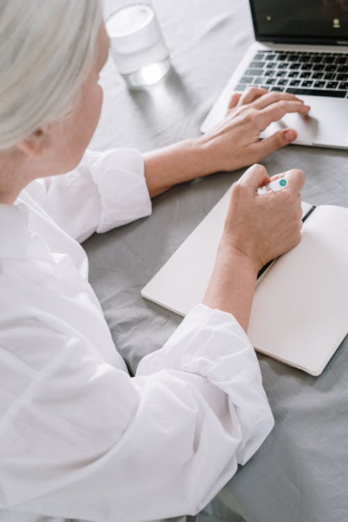 An Elderly Woman Taking Notes While Using A Laptop