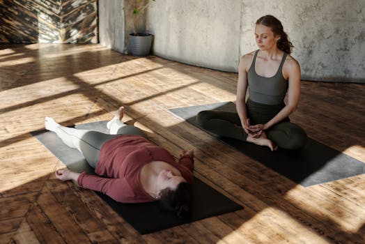 Two women practicing yoga indoors, one guiding the other in a relaxing pose on a wooden floor.