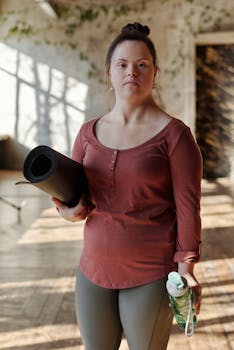 A young woman preparing for a yoga session indoors, holding a mat and water bottle.