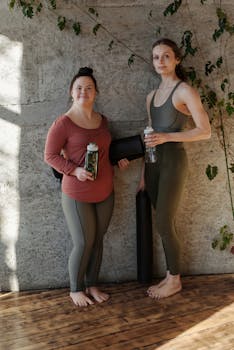 Two women holding yoga mats and water bottles, ready for a yoga session indoors.