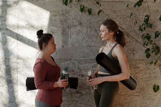 Two women chatting indoors after a yoga session, each holding a water bottle and yoga mat.