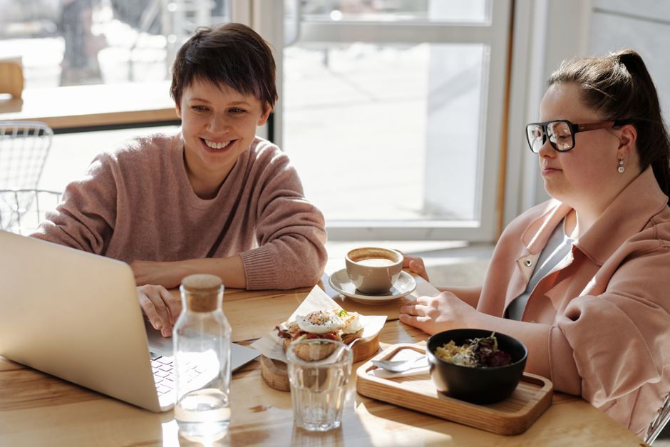 Two women enjoying coffee and snacks while working on a laptop in a bright café setting.
