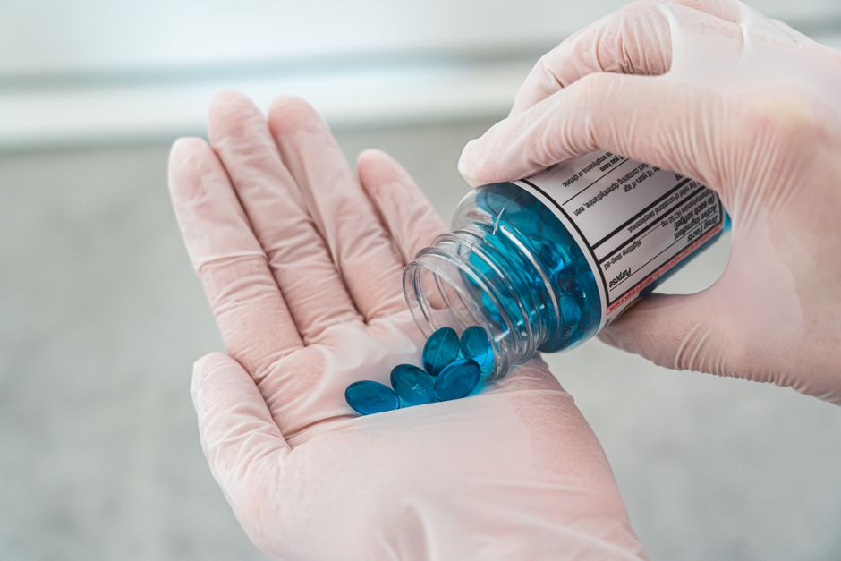 Gloved hands carefully pouring blue oval pills from a prescription bottle.