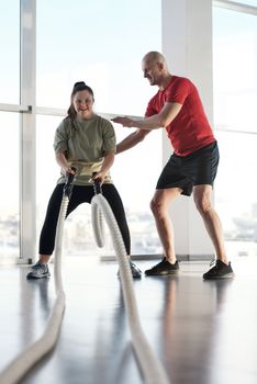 A woman with Down syndrome working out with a trainer using battle ropes in a gym.