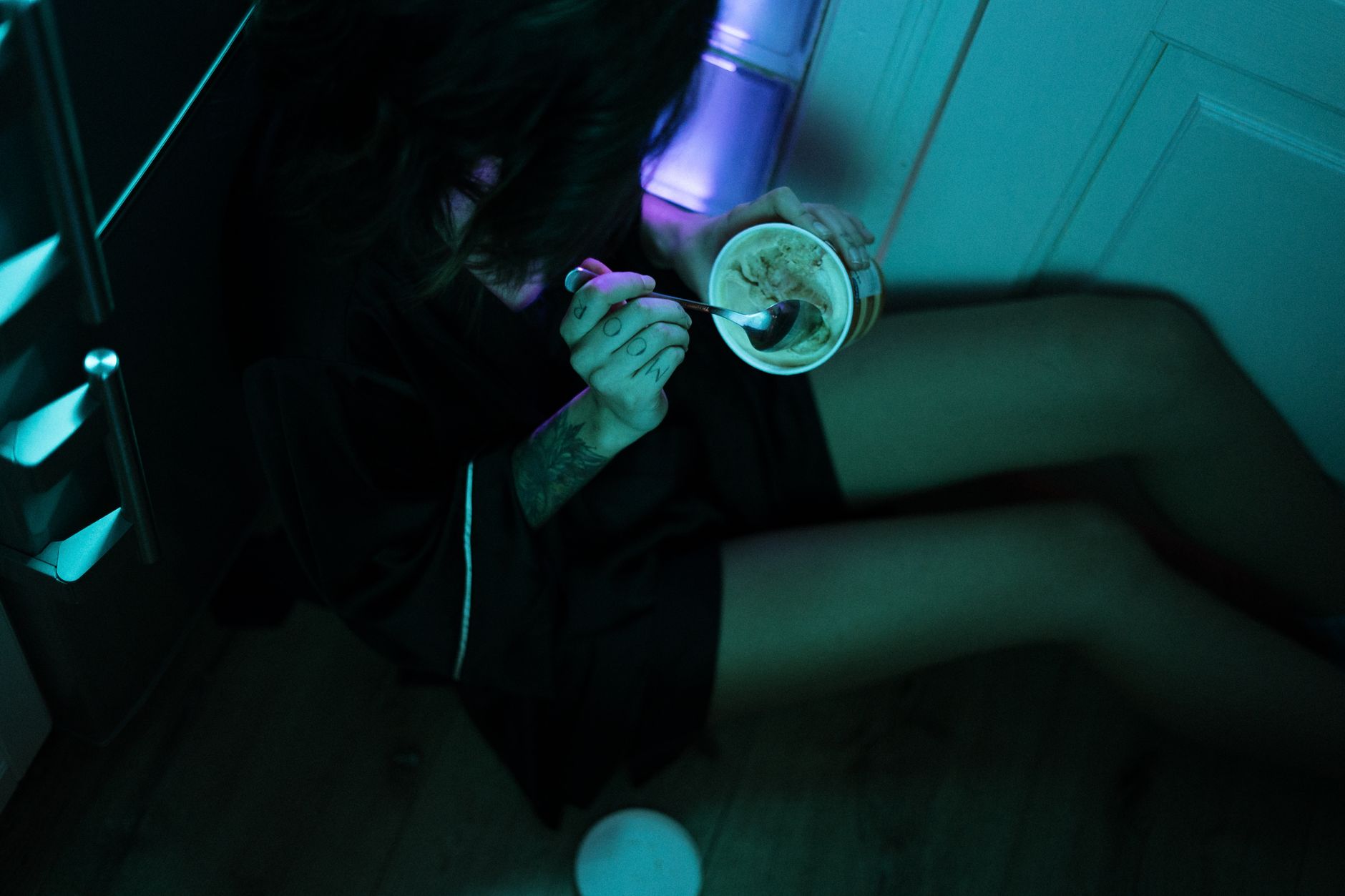 A woman enjoys ice cream on the floor of a dimly lit kitchen with neon lights, conveying isolation.