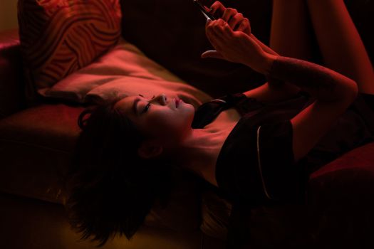 Asian woman relaxing with her smartphone in a cozy home setting, under atmospheric lighting.