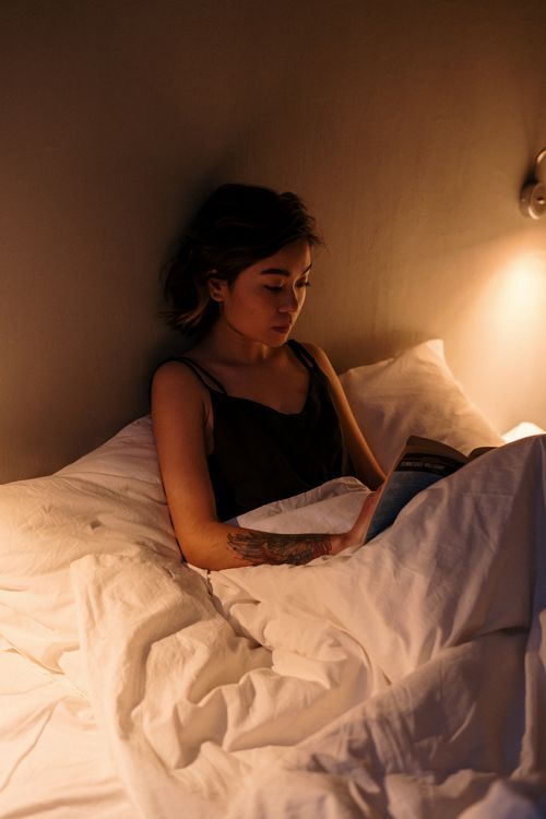 Free Woman enjoys a quiet evening reading in bed under soft lighting and cozy blankets. Stock Photo