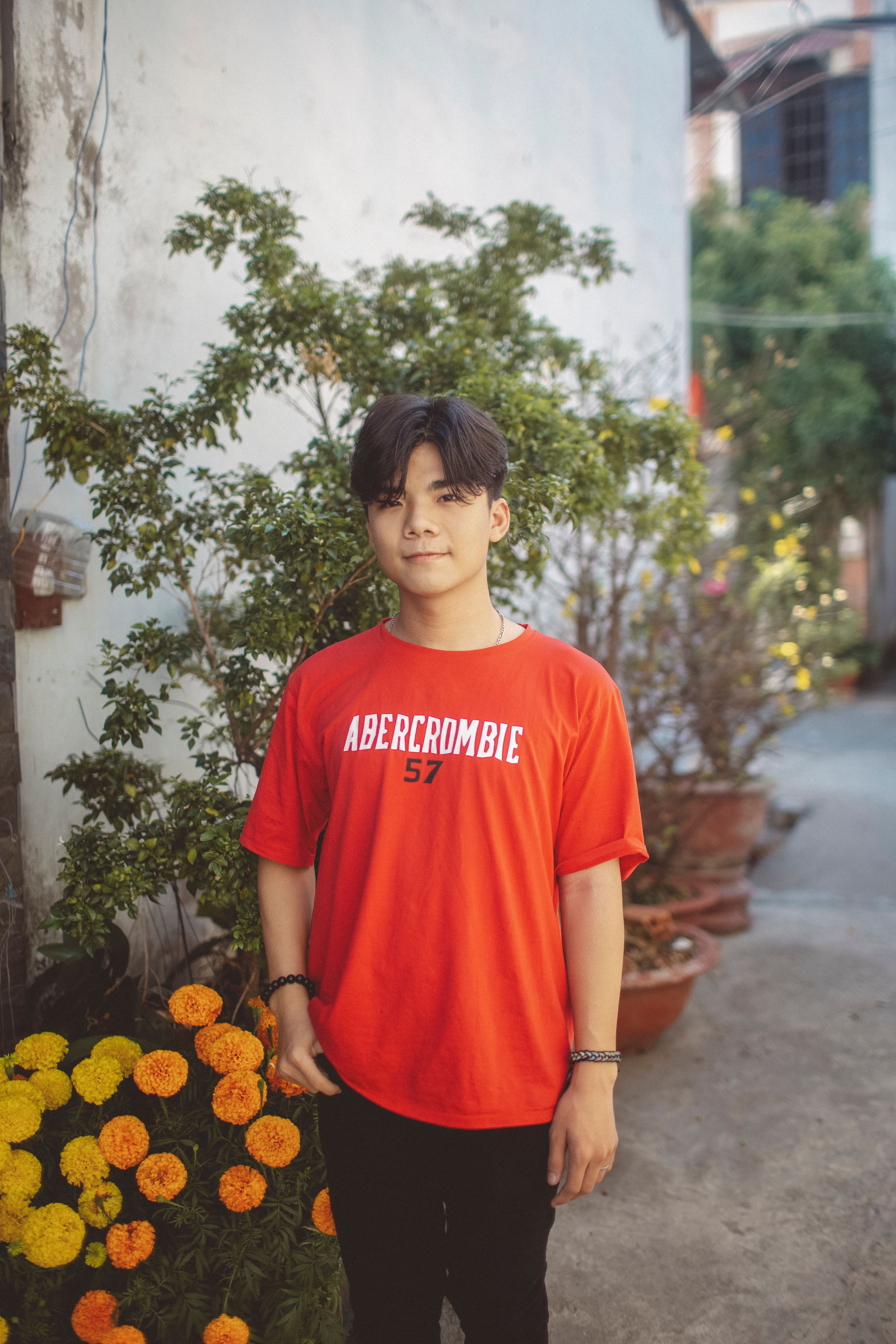 Smiling young man in a red shirt stands casually outdoors with potted plants.