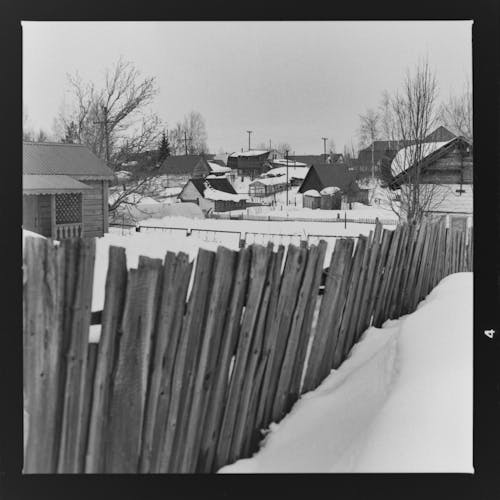 Black and white shot wooden fence tilting near heap of snow against aged shacks on winter day in village
