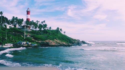 Striped lighthouse located on coast with tropical plants near stormy sea against cloudy sundown sky