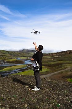 A man controls a drone in a vast, scenic landscape with a winding river and green hills under a blue sky.