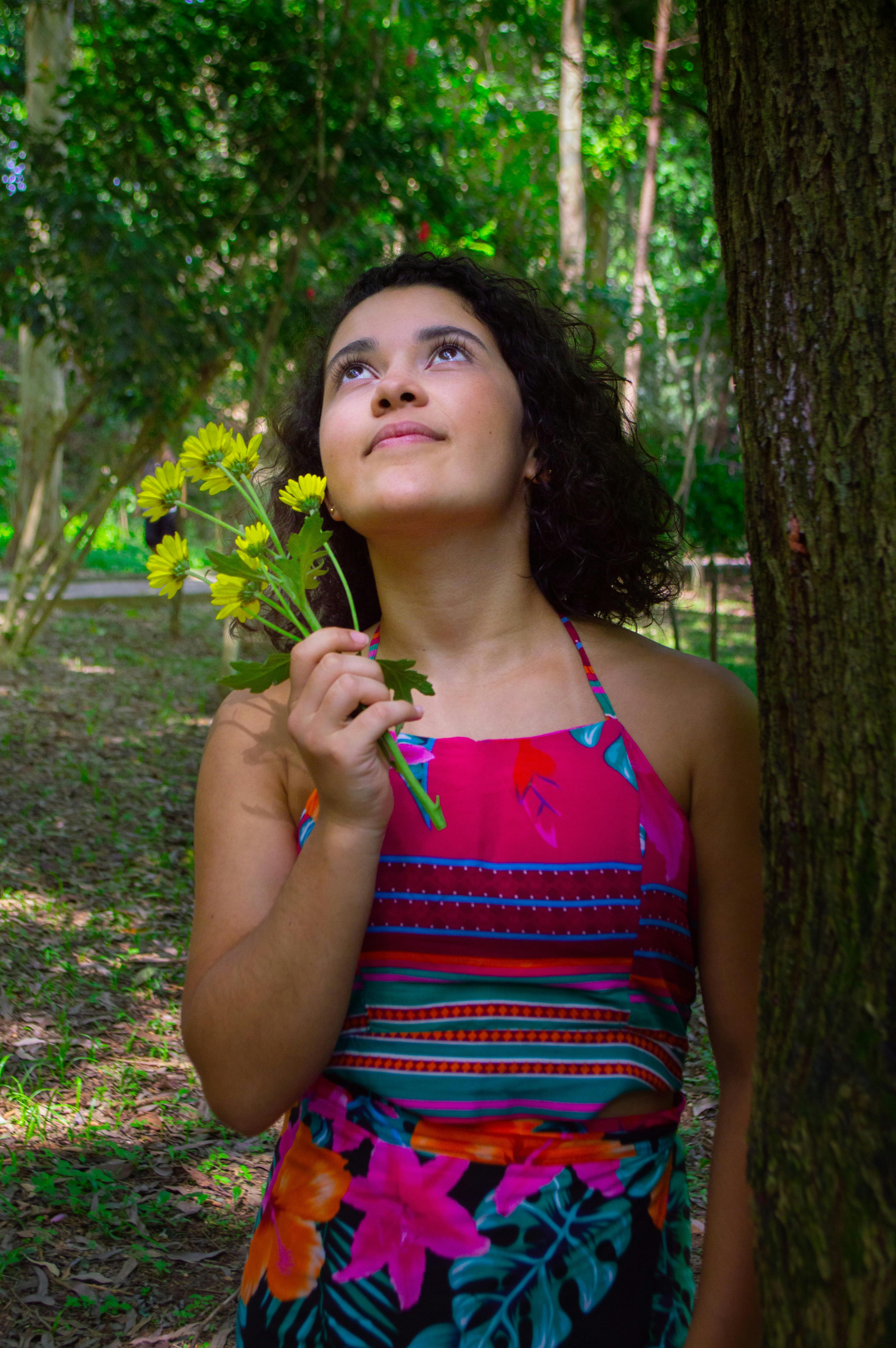 Woman in a vibrant dress holding flowers, looking upwards in a lush outdoor setting.