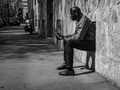 Free Side view of African American guy browsing smartphone and listening to music while sitting in hole in building wall on city street Stock Photo