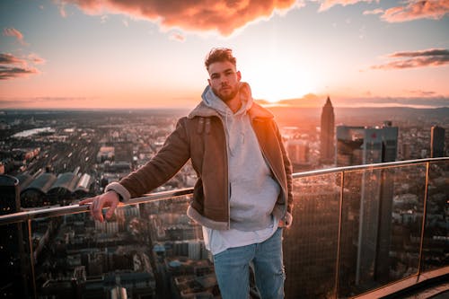 Free Man in Brown Zip Up Jacket and Blue Denim Jeans Standing on Top of Building during Stock Photo