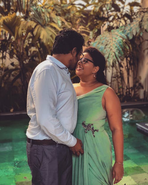 Adult Indian man and woman in elegant clothes looking at each other and smiling while standing near pool during date in tropical garden