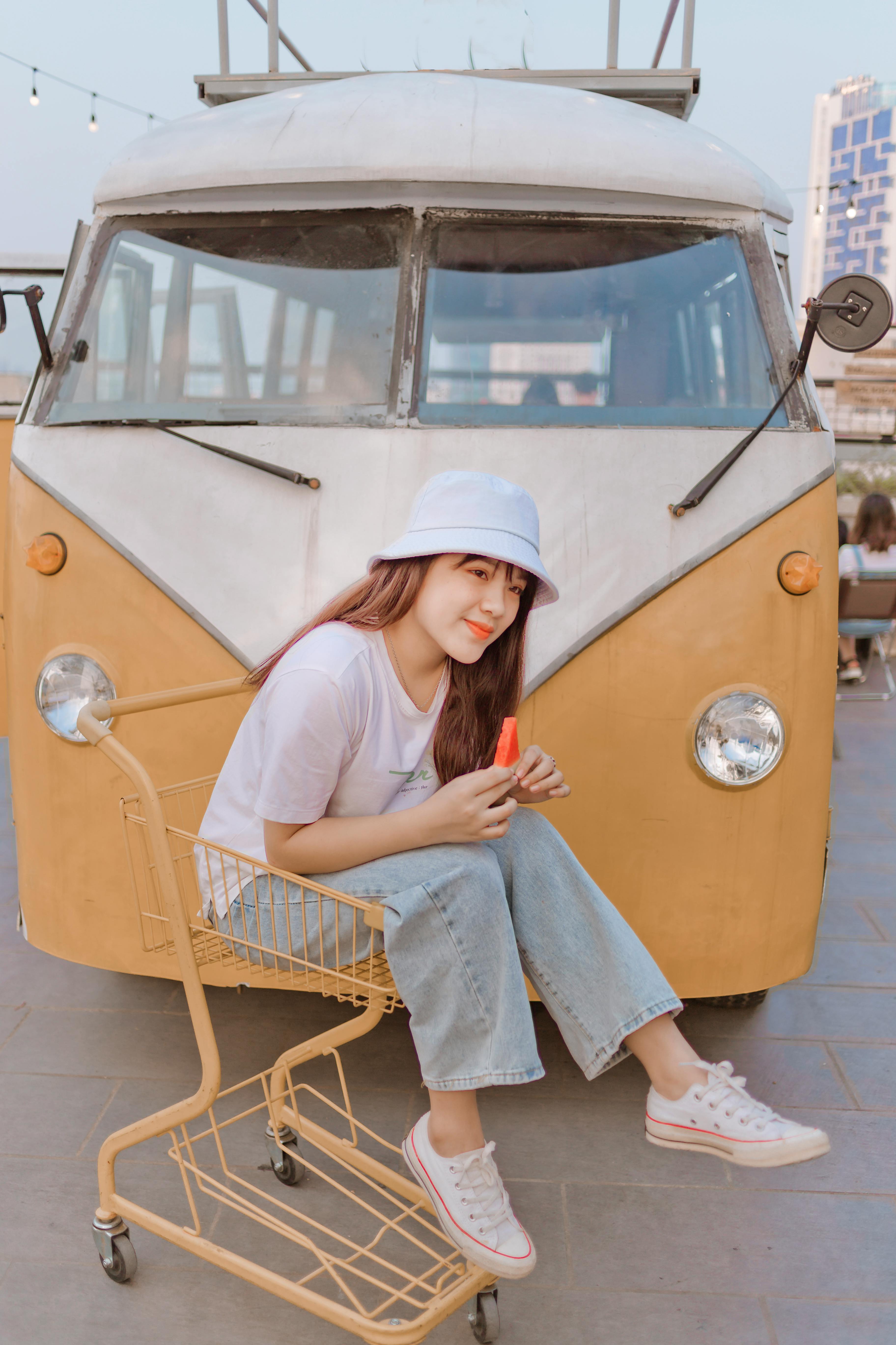 Cheerful young Asian female in casual clothes sitting in shopping trolley and enjoying fresh watermelon near retro van
