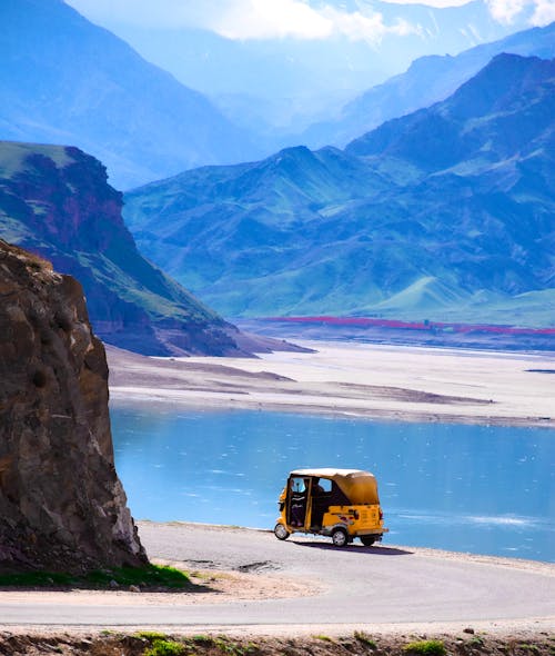 Yellow car driving on curvy road near calm lake with blue water and mountain ridge on sunny day in nature