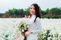 夏日田野里，一位身着白色连衣裙的快乐年轻亚洲女性手握鲜花，对着镜头微笑