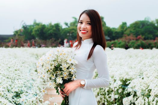 夏日田野里，一位身着白色连衣裙的快乐年轻亚洲女性手握鲜花，对着镜头微笑