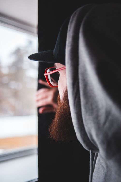Free Side view of bearded male hipster in hoodie and glasses looking out window in cozy room at home Stock Photo
