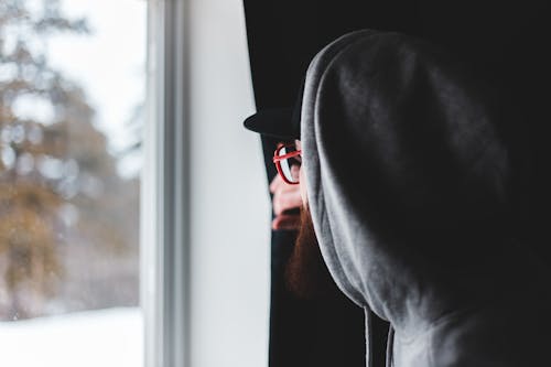 Side view of bearded man in glasses and hoodie pulling curtain and looking out window on winter day at home