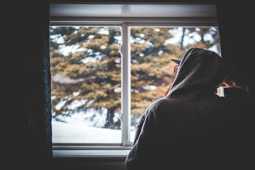 Back view of unrecognizable guy in hoodie and glasses resting near window and admiring snowy weather on winter day at home