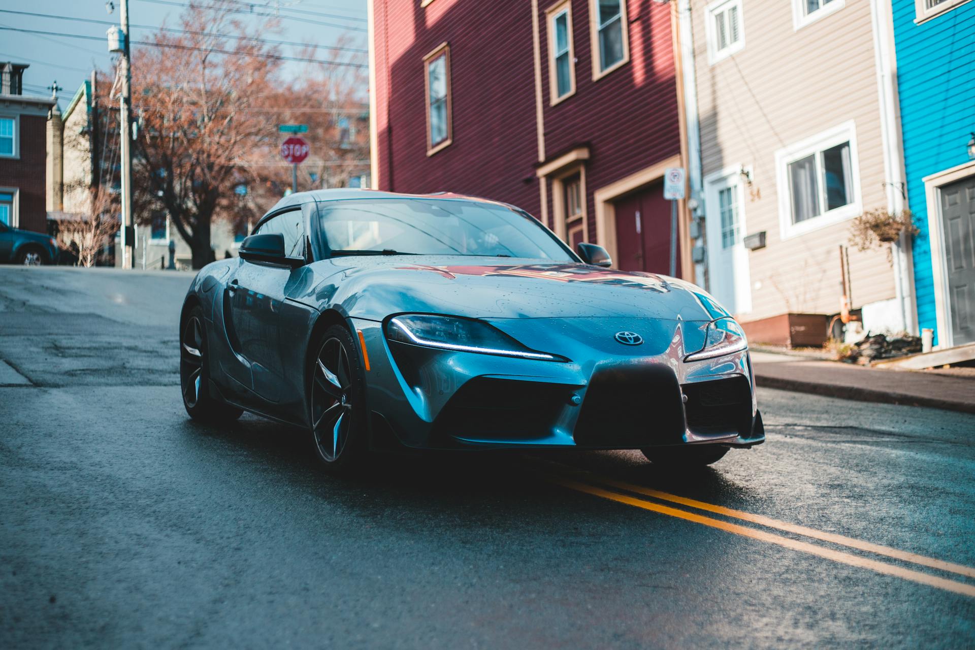 A blue Toyota Supra parked on a wet city street, showcasing its elegant design against colorful houses.