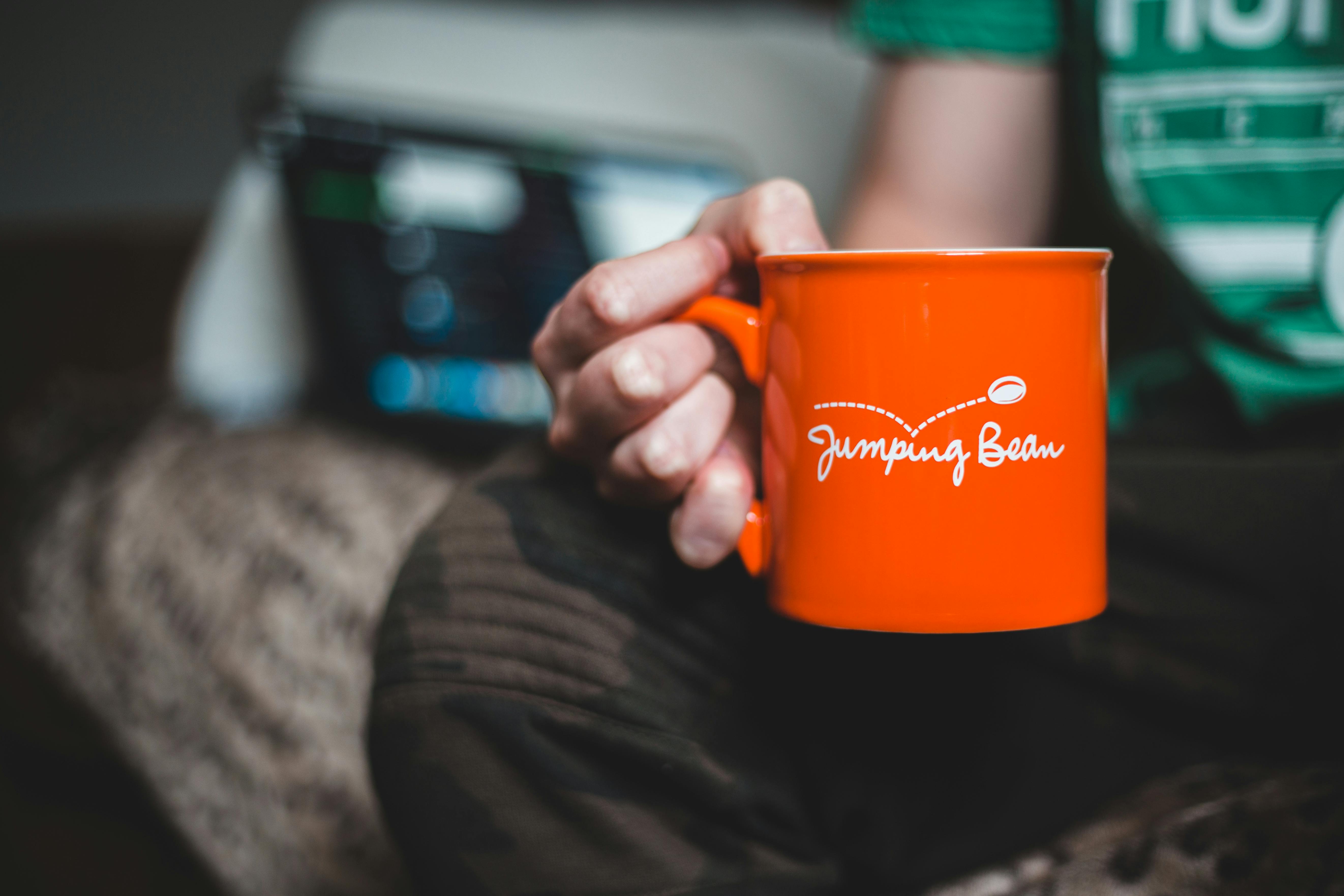 Close-up of a person holding an orange mug with 'Jumping Bean' text indoors.