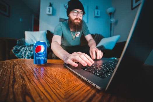Bearded man wearing beanie, typing on laptop with soda can in cozy workspace.
