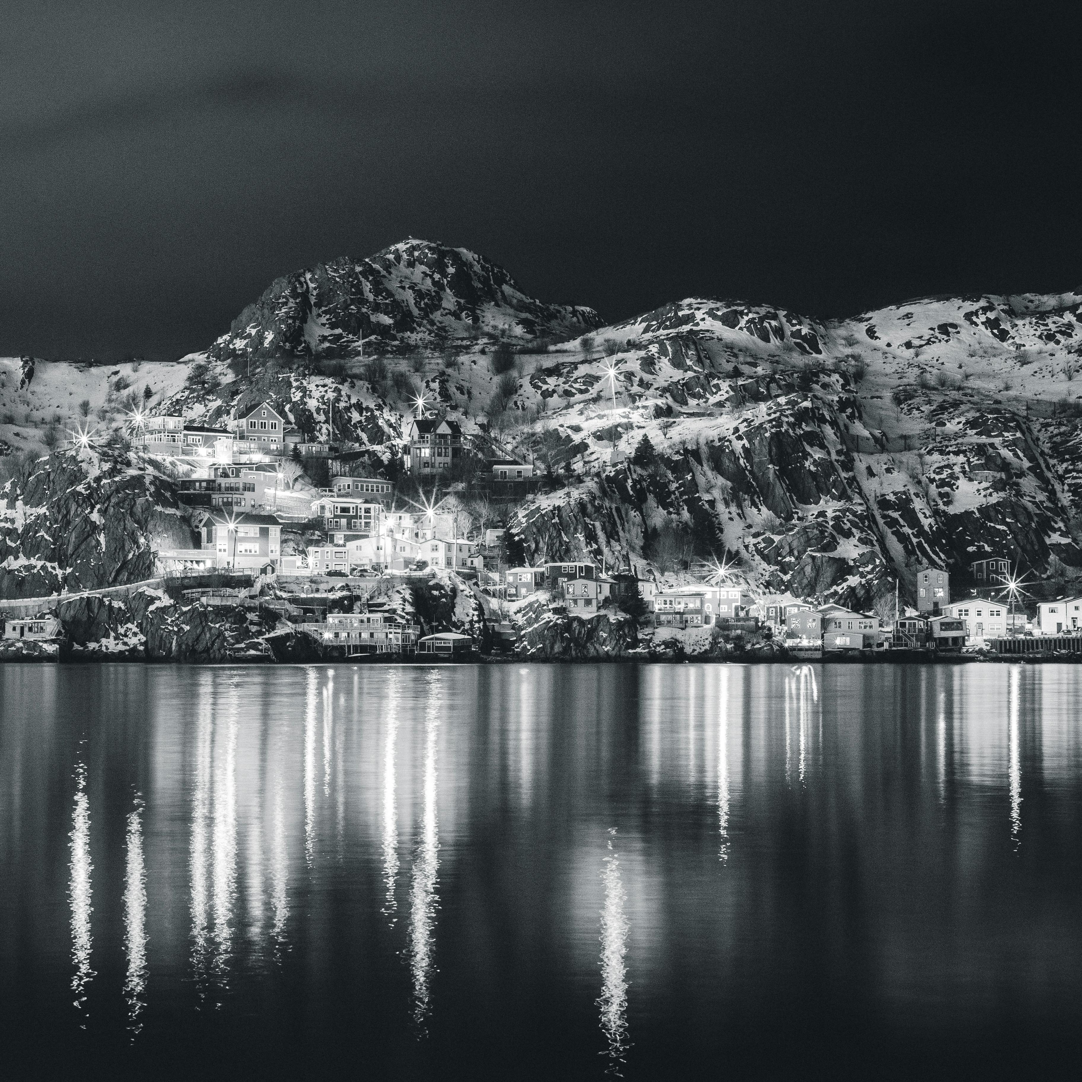 Black and white glowing town on shore of peaceful bay with mountains reflecting in dark water at night