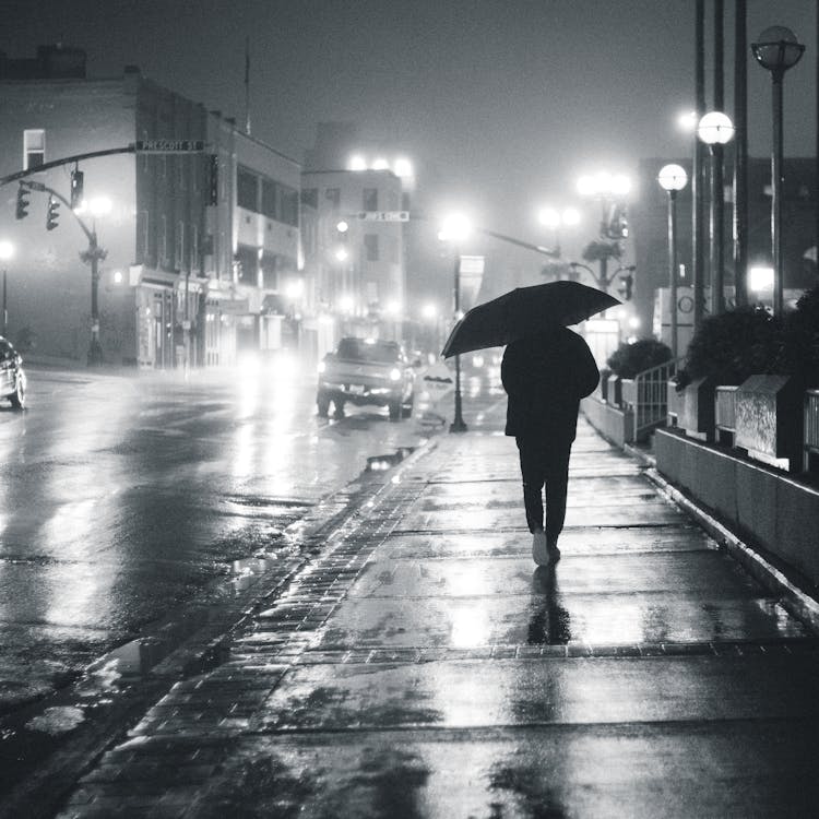Silhouette of person with umbrella walking on wet sidewalk with glowing lamps and cars at night