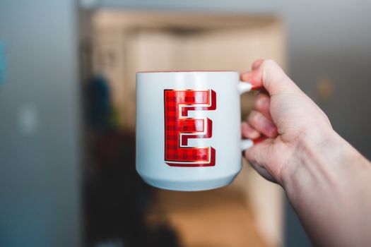 Hand of person holding white ceramic cup with tartan letter E at home
