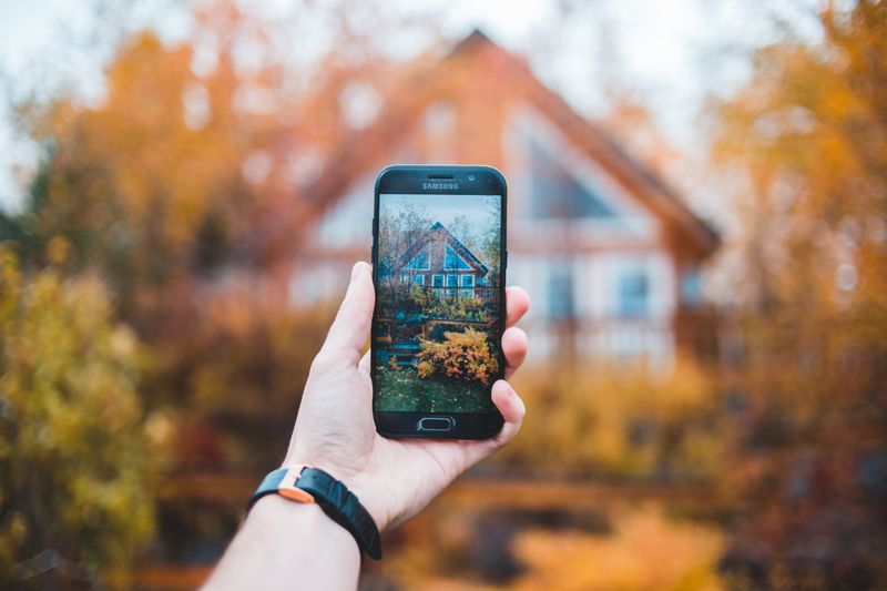 Hand holding smartphone photographing a residential property exterior with autumn foliage