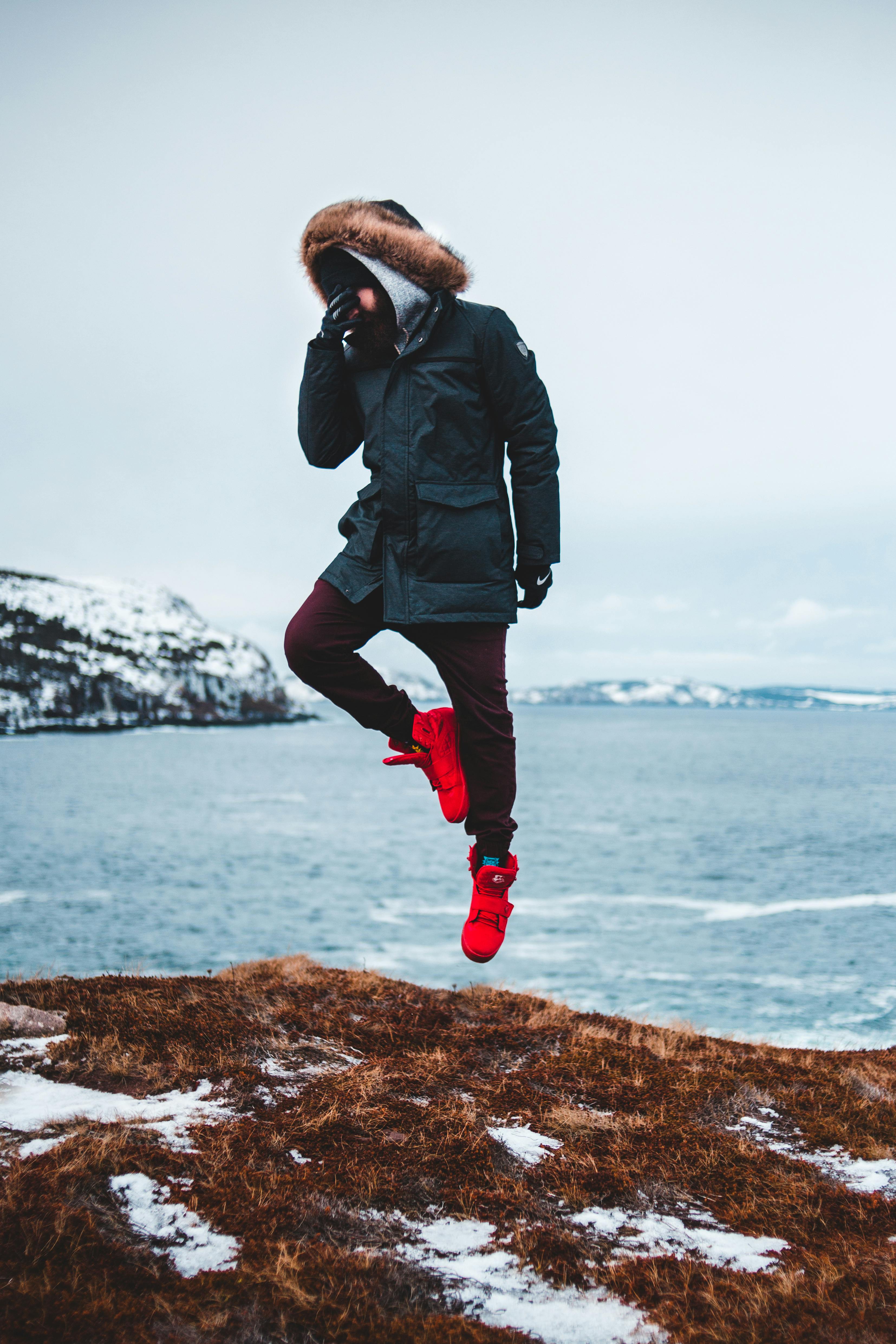 A person in winter attire jumps joyously by the snowy ocean cliff.