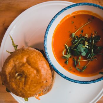 Top view of appetizing burger with crunchy golden surface on plate near bowl with cream soup decorated with fresh watercress leaves on table in cafeteria