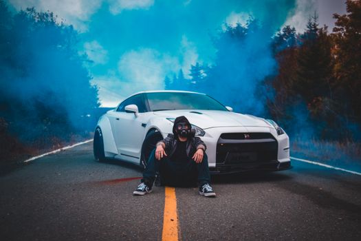 Anonymous male in trendy wear and respirator sitting on asphalt roadway near contemporary auto among trees with smoke behind in evening