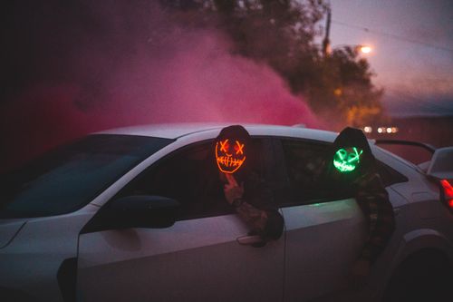 Free Anonymous male friends in fearful shining masks driving modern auto on city street behind trees and smoke in air in evening illuminated by artificial light Stock Photo