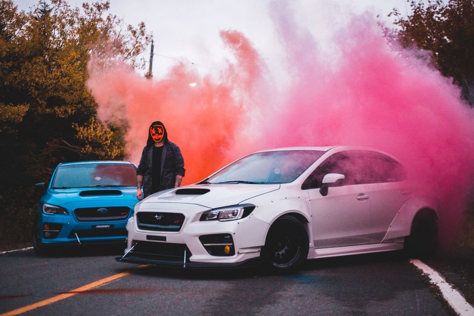 Unrecognizable hipster male in spooky mask standing on roadway near contemporary automobiles with colored smoke behind in countryside