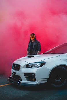 Masked man stands beside a white sports car enveloped in colorful smoke, creating a mysterious and vibrant urban scene.