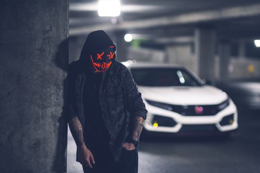 Anonymous tattooed male in trendy wear and neon mask standing with hand in pocket near modern auto on floor in parking garage illuminated by ceiling lamp