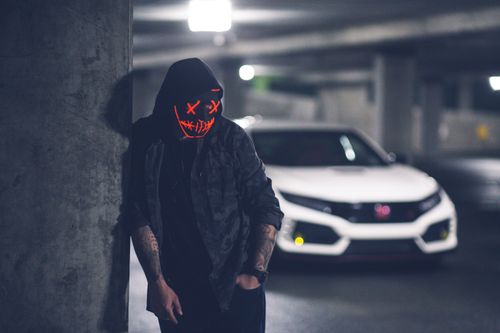 Free Anonymous tattooed male in trendy wear and neon mask standing with hand in pocket near modern auto on floor in parking garage illuminated by ceiling lamp Stock Photo