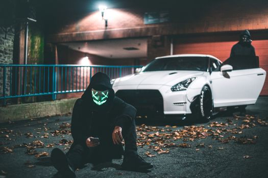 Anonymous male in neon mask browsing internet on cellphone while sitting on asphalt road covered with faded leaves near modern auto and friend standing on illuminated street at night