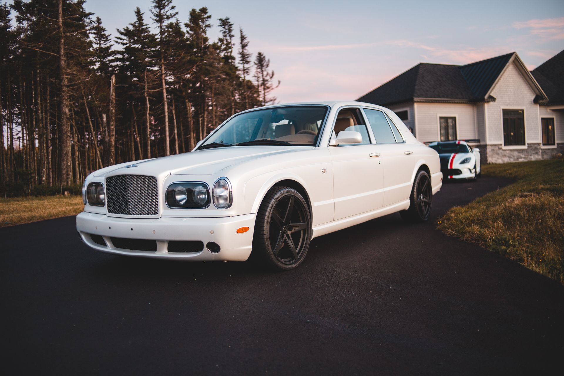 A classic luxury white car parked on a suburban driveway beside a modern house.