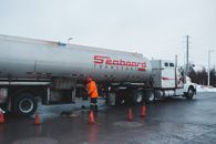Back view of anonymous male worker in bright uniform and helmet cleaning oil truck vehicle while standing on asphalt roadway surrounded by barriers under white sky in town
