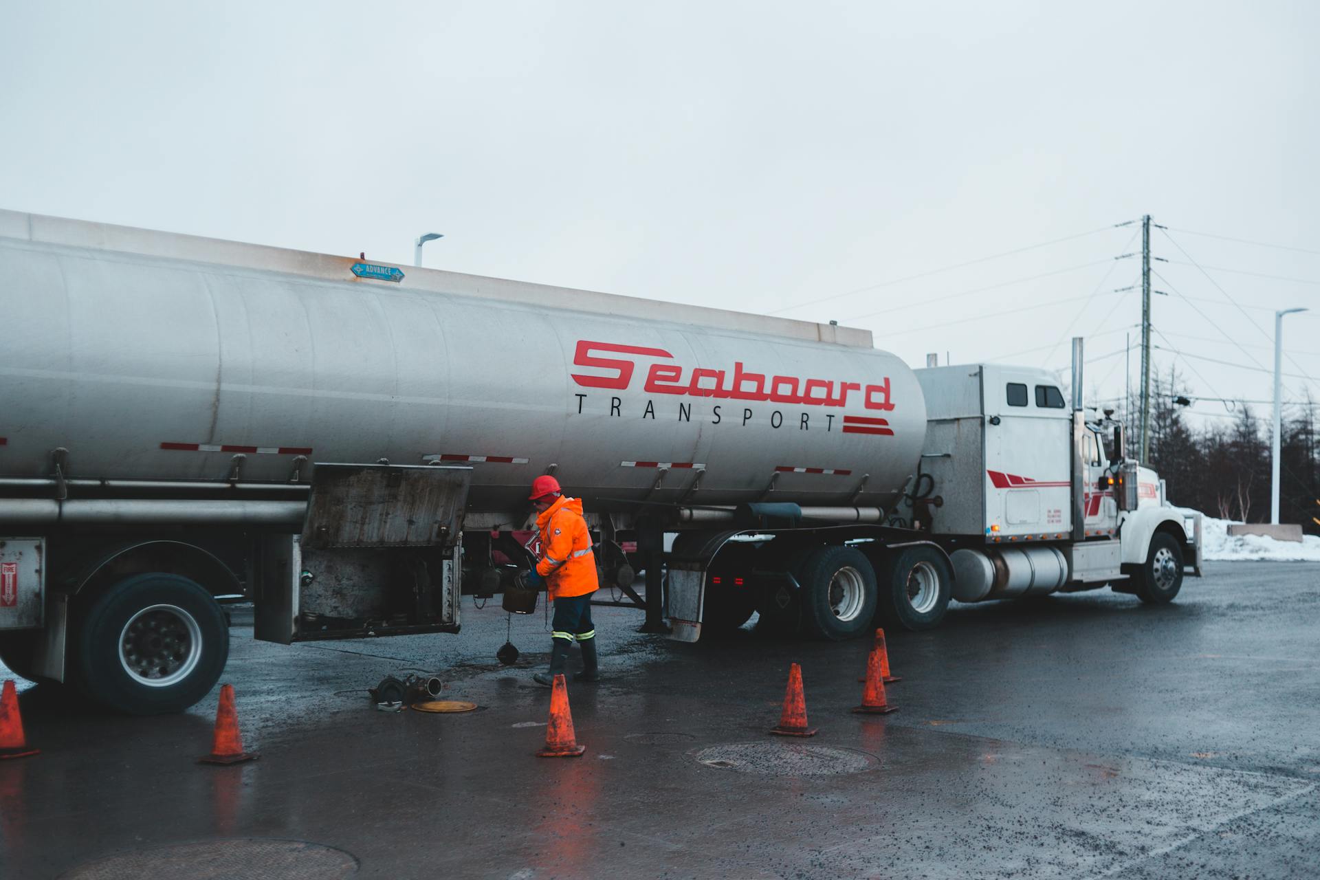 Back view of anonymous male worker in bright uniform and helmet cleaning oil truck vehicle while standing on asphalt roadway surrounded by barriers under white sky in town