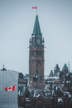 A low angle shot of the Peace Tower with snow and a Canada flag flying, highlighting Gothic architecture.